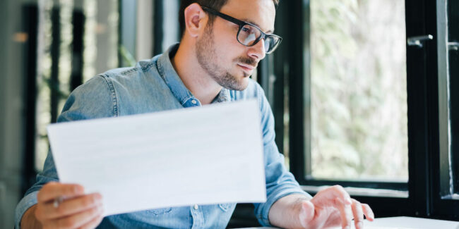 Serious pensive thoughtful focused young casual business accountant bookkeeper in office looking at and working with laptop and income tax return papers and documents